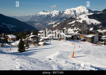 A sunlit ski slope with snow making equipment and a ski resort in the background set in the snow covered Alps, under blue skies. Stock Photo