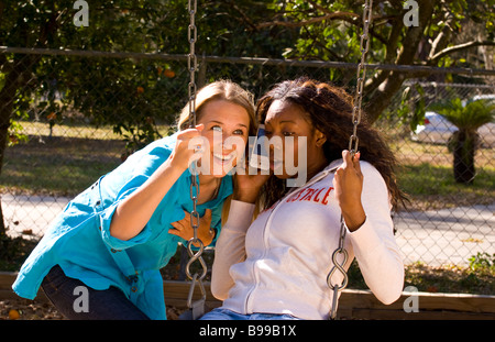 Two young women aged 20s playing with on swing outdoors in park laughing and having fun playing with Iphone listening Stock Photo