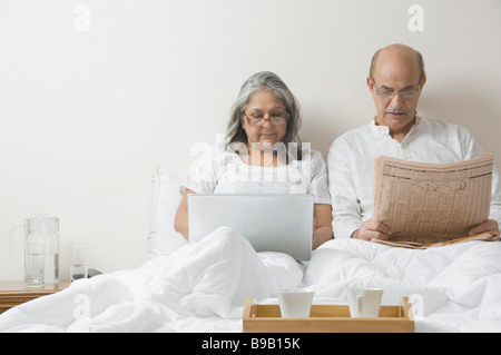 Man reading a newspaper with a woman using a laptop on the bed Stock Photo