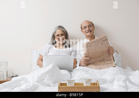 Man reading a newspaper with a woman using a laptop on the bed Stock Photo