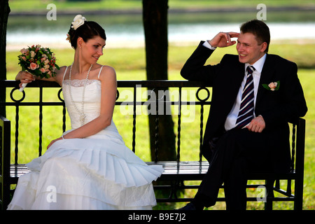 Newlywed couple sat on park bench smiling groom and coy bride with bouquet Stock Photo