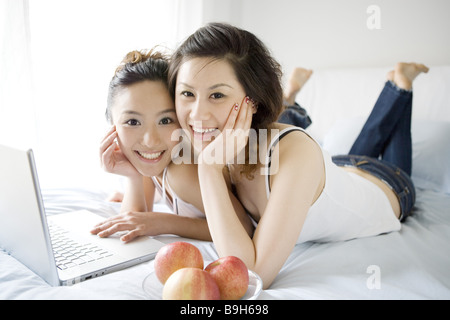 Two young women lying on bed with laptop and apples smiling portrait Stock Photo