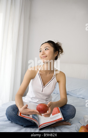 One young woman sitting on bed with magazine and apples Stock Photo