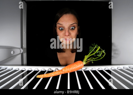 Young woman regarding a single carrot in an empty refrigerator