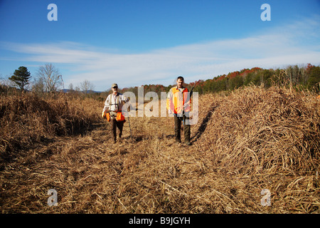 A hunting guide leads a bird hunter orange vest and gun through an open field in search of game