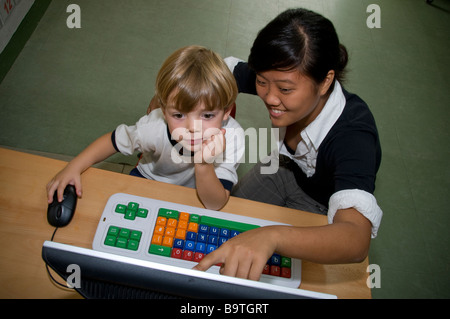 Infant boy 4-6 years in kindergarten class working with mouse on his colour coded computer with help from his young female school computer teacher Stock Photo
