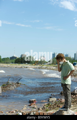 Boy standing on polluted shore, wearing pollution mask, head down Stock Photo