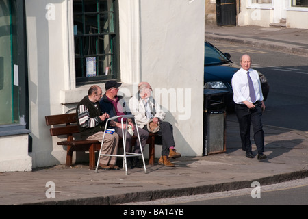 Three pensioners sitting on a street corner bench observing a passing business man Stock Photo