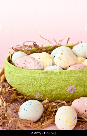 Pastel-coloured mini Easter eggs in a papier mâché egg-shaped container with straw, on a plain pale pink background Stock Photo