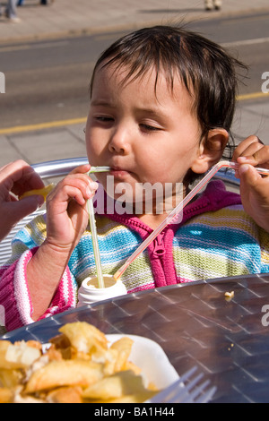 Young girl on her summer holidays drinks from a milk shake bottle using two straws with a plate of chips waiting to be eaten Stock Photo