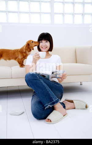 Young woman sitting on floor holding cup and book Dachshund licking her face on sofa behind Stock Photo