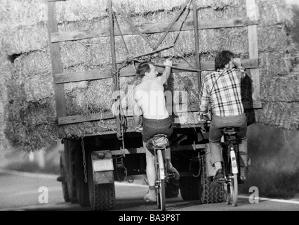 Seventies, black and white photo, autumn, hay harvest, tractor brings in the harvest, two cyclists hang on the trailer, men, aged 30 to 40 years Stock Photo