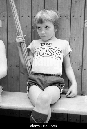 Seventies, black and white photo, edification, school, sports, physical education, gym class, gymnastics, sports hall, little boy sits on a bench and holds to a thick rope, waiting for his appearance, inscription on his tricot The Boss, aged 5 to 8 years Stock Photo