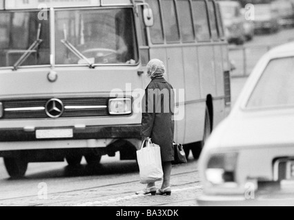 Seventies, black and white photo, people, older woman with shopping bags stands in the middle of a traffic road and tries to cross the road, heavy traffic, aged 60 to 80 years Stock Photo