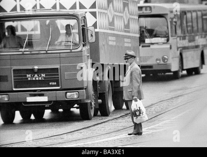 Seventies, black and white photo, people, older man with shopping bag stands in the middle of a traffic road and tries to cross the road, heavy traffic, aged 65 to 75 years Stock Photo