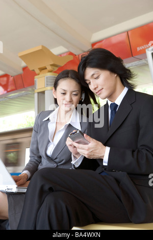 Businessman sitting on bench showing cell phone to a businesswoman on train platform Stock Photo