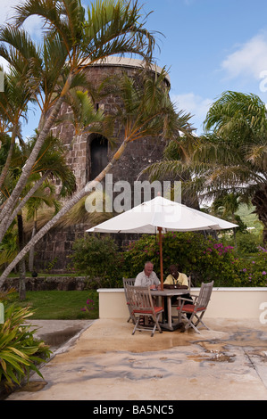 Two men sitting at a table table under an umbrella at Montpellier Plantation Inn with a disused sugar mill behind Stock Photo