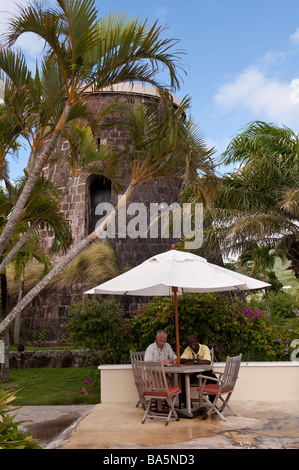 Two men sitting at a table table under an umbrella at Montpellier Plantation Inn with a disused sugar mill behind Stock Photo