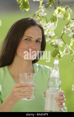 Woman long-haired brunette mineral water bottle glass holds gaze camera spring outside portrait 30-40 years garden tree blooms Stock Photo