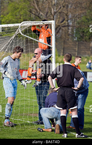 A footballer sits on the shoulders of the groundsman in order to mend a net which broke during the match whilst the ref looks on Stock Photo