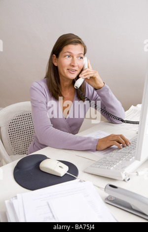 Studies desk woman works computers telephones smiles semi-portrait apartment office people 40-50 years independent freelance Stock Photo