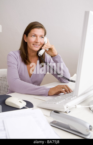 Studies desk woman works computers telephones smiles semi-portrait apartment office people 40-50 years independent freelance Stock Photo