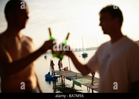 Two men drinking bottles of beer, children playing on jetty in background, Lake Woerthsee, Bavaria, Germany, MR Stock Photo
