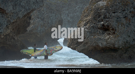 Two surfers hold their surfboards and check out the waves between rocks before entering the sea to surf Stock Photo