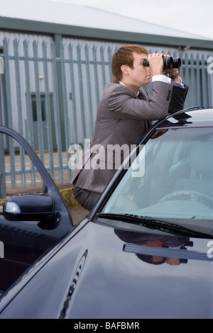 Young man leaning on car looking through binoculars Stock Photo