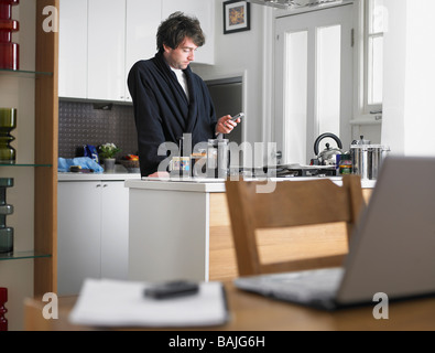 Man standing in kitchen, text messaging, laptop on table in foreground Stock Photo