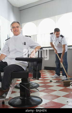 Two barbers in barber shop, one sweeping floor Stock Photo