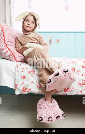 Young girl (5-6) sitting on bed wearing bunny costume and monster slippers Stock Photo