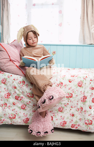 Young girl (5-6) sitting on bed wearing bunny costume and monster slippers, reading book Stock Photo