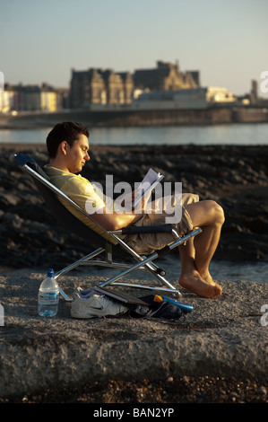 A young male university student sitting in chair on the beach at Aberystwyth reading a text book in the sunshine Wales UK Stock Photo