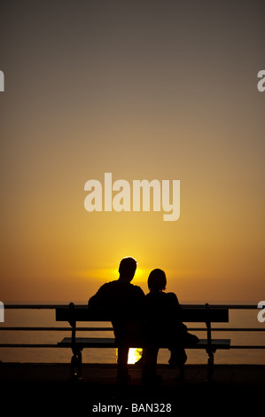A romantic young couple sitting on a seaside bench silhouetted against the sunset on a warm summer evening Aberystwyth wales UK Stock Photo