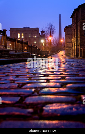 A view down the cobbled street of Albert Terrace, with a view of Salts Mill and its chimney, Saltaire, Bradford, West Yorkshire Stock Photo
