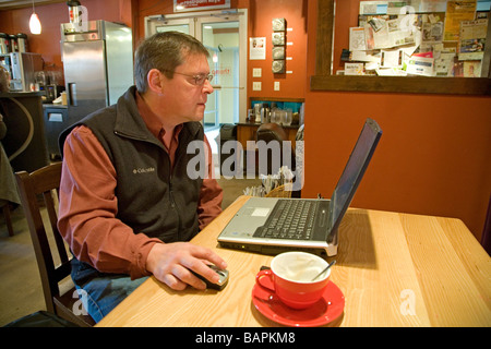 A man works on his lap top computer and drinks a cappuccino coffee at a coffee house in a downtown business area Stock Photo