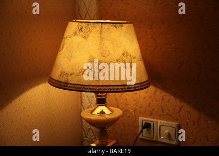 it's a lamp in the corner of a room in a hotel. It is plugged and the light is on. Color is yellow and brown. Inside. Old style. Stock Photo