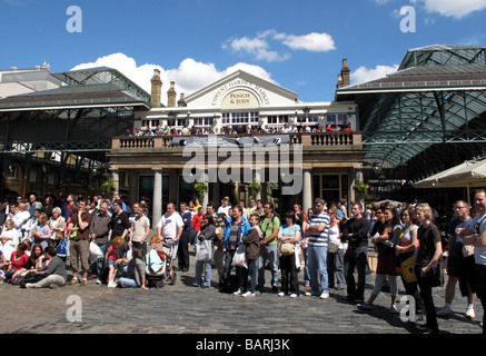 Crowds of tourists watching street artists busking in the sunshine outside Punch and Judy pub at Covent Garden Market in London. Stock Photo
