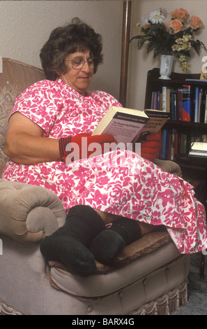 elderly woman sitting indoors reading a book with gloves and socks on due to poor circulation Stock Photo