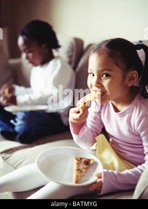 Junk food. Five-year-old girl taking a bite from a slice of pizza. In the background, a five-year-old boy is also eating pizza. Stock Photo