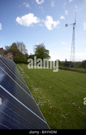 sun shining down on an array of blue tinted polycrystalline silicon photovoltaic solar panels and medium sized wind turbine Stock Photo