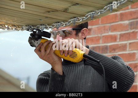 Young man wearing protective eye gear using small angle grinder on chain-link bridge. Stock Photo