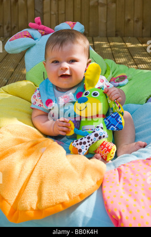 Vertical close up portrait of a cute six month old baby girl sitting on a colourful cushion in the garden on a sunny day Stock Photo