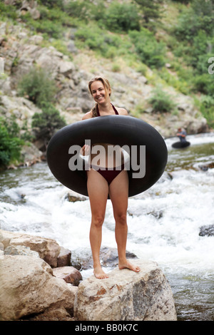 A young caucasian woman in a bikini stands next to a river with a large inner tube. Stock Photo