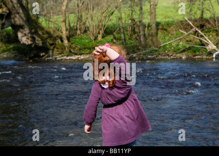 Little girl with red curly hair throwing stones into the river on a sunny day. Stock Photo