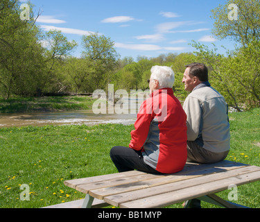 senior couple sitting on picnic table looking at river on spring day Stock Photo