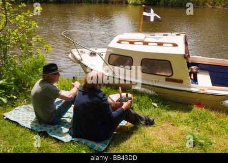 A middle aged couple enjoying a picnic at the riverside next to their boat on the river Waveney on the Norfolk Broads in the uk Stock Photo