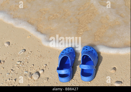 Pair of blue beach shoes or crocs at the edge of the sea with a small wave and sand Stock Photo