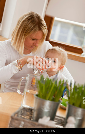 Mother helping her 1-year-old daughter to drink water from a glass Stock Photo